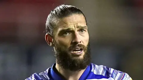 PA Media Andy Carroll during a game. He is wearing a blue and white-striped football kit. He has his black hair pulled back into a bun and has a bushy dark beard. He is sweaty during a football match.