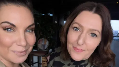 Hayley Turner Two women are pictured posing for a selfie. Hayley has her hair scraped back and she is wearing a trench coat, Jo has shoulder-length hair and is wearing a green and black dress.