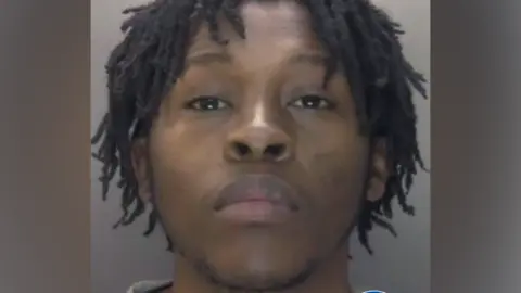 A police custody photo of Koketso Ximba, who is wearing a grey top and is in front of a grey wall