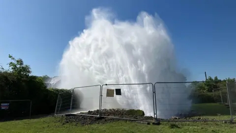 A huge spurt of water shoots into the sky, behind a temporary metal fence. The earth around the explosion has been thrown up with the force of the burst.