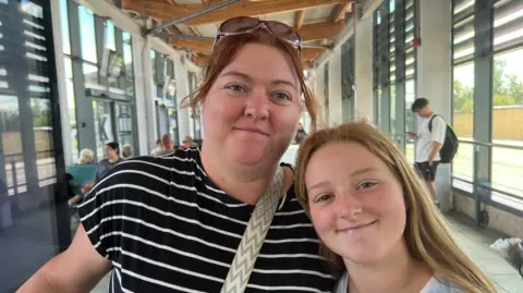A close up shot of a mother standing with her her daughter in a modern, glass-fronted bus station. The mother has reddish-brown hair, tied back, with sunglasses on the top of her head and is wearing a black T-shirt with white stripes. Her daughter has long strawberry blond hair, is wearing a white top and is smiling.