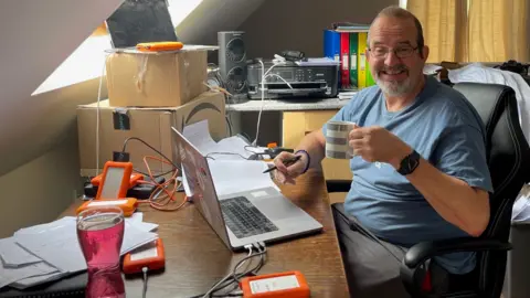 Tread The Globe A man in a blue t-shirt with a mug of tea sat at a desk with a laptop and several orange hard drives