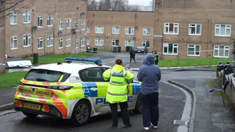 A police car parked in the middle of a residential street. A police officer is standing next to the car, in a high-vis jacket, with another person standing beside them. The other person is wearing a blue hoodie. There are houses built from yellow brick in the background, with windows on the ground and first floor.