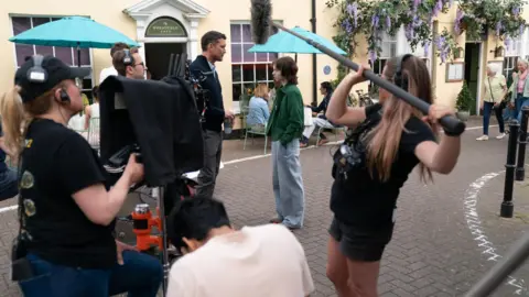 Somerset Council A woman uses a film camera wearing a hat and earphones, and next to her is a woman in black holding a boom microphone. They are on a pretty street with houses covered in wisteria. In the middle of the shot are a man and woman standing looking at each other, as if in conversation.