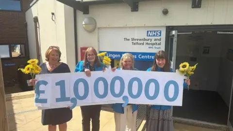 Four women are standing in a line and smiling. They are holding bunches of yellow flowers and a line of blue numbers on paper. The numbers read "one million pounds"