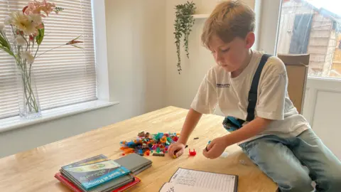 Austin sat on a wooden table which has books and LEGO blocks in a pile.