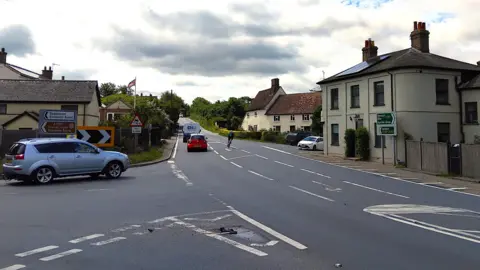 A crossroads at Earl Stonham in Suffolk. There are houses on either side. The A140 is running through the middle of the picture. Cars are waiting to join it from both sides of the crossroads.