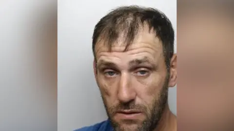 A mugshot of a man in his forties. He has thin, short, dark hair, sunken eyes and brown stubble.