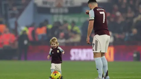 Reuters A toddler in an Aston Village football kit has his mouth opened excitedly while kicking a football. An Aston Villa player with his back to the camera can be seen pointing. In the background a stadium full of fans can be seen