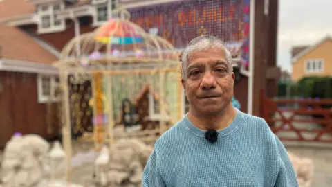 Jagdish Gohil is pictured outside his house which has been decorated in hundreds of lights for Diwali. He is wearing a blue jumper and has short grey hair. 