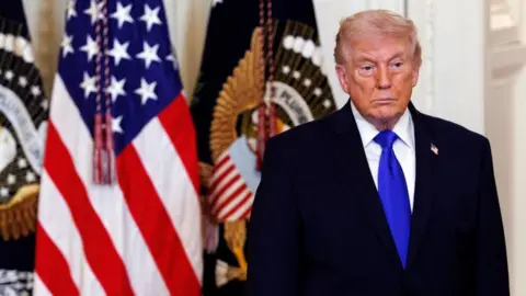 US President Donald Trump stands expressionless in front of US flags at the White House
