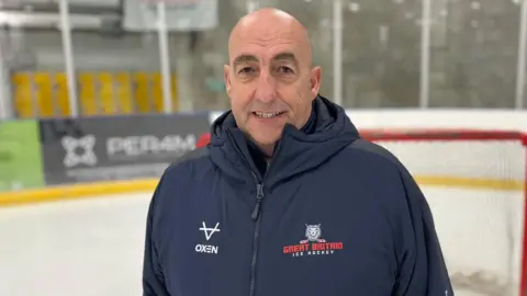 A bald headed man in a dark blue Great Britain ice hockey jacket