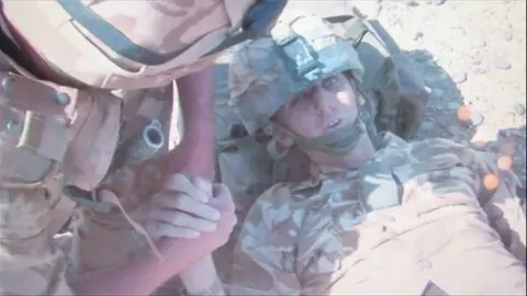 Johno Lee Two soldiers in desert camo army gear. One solider who's face is not in shot is holding the hand of a another solider Johno Lee lying on the floor who has been injured in IED blast. His head is resting on his backpack.