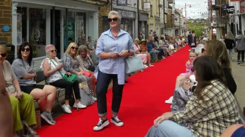 Discover Daventry A woman with short white hair and dark glasses poses on a red carpet in a shopping street, wearing a blue shirt, light-coloured handbag, denim trousers and blue and white plimsolls. There are spectators sitting on both sides of the carpet, and shops behind them.