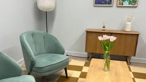 Linc Charity Two green armchairs are arranged around a wooden coffee table with a vase holding pink tulips. The coffee table is on a beige and brown checkerboard rug. There is a sidetable with decorations on and two paintings above. There is a white lamp in the corner. The walls are painted an off-white colour.