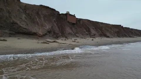 Waves are seen in the foreground and beyond that the tall brown cliffs where the brown brick bunker is perched.