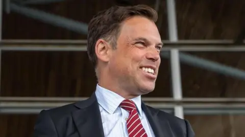Ken Skates smiling off to the side of the image, wearing a dark suit and red and white tie, with a pin stripe shirt.
