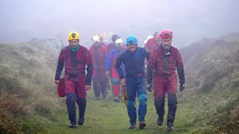 PA Media Several members of the rescue crew walking above ground following their efforts underground to help George Linnane. The air is misty. They are wearing safety helmets and workwear overalls.