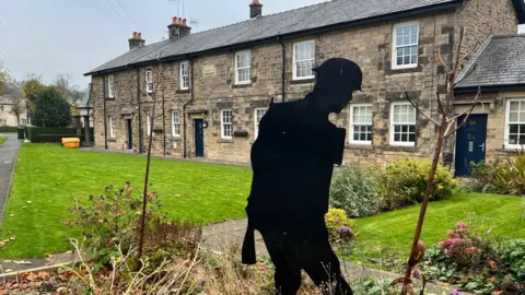 BBC/Paul O'Gorman General view of the village showing a row of stone cottages behind a rectangular patch of neat grass. There is a black silhouette of a soldier in the foreground