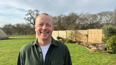 Daniel Adams is smiling. He is wearing a green over-shirt. There is green grass and a fence behind him. He is standing on a farm and you can see trees and a blue sky.