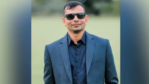 Ketan Shah smiling for a picture outside. He is wearing a suit and shades.