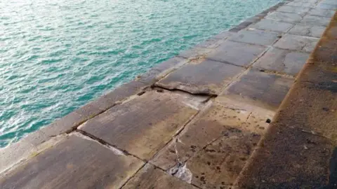 States of Guernsey The breakwater stretches foward into the turqose sea, with a damaged area seen in the foreground. 