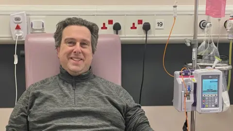 Phil Grisewood A man in a charcoal grey quarter zip top sits in a chair in a hospital. There are pieces of medical equipment surrounding him. 