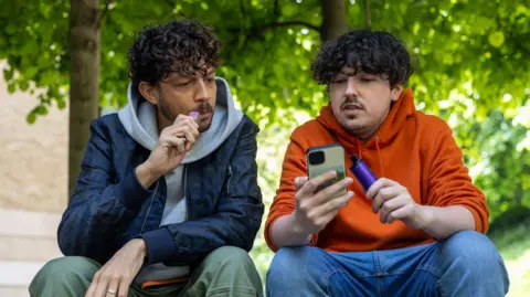 Two young men sit outside vaping. They are chatting, while looking at a mobile phone. 