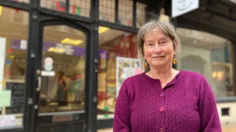 Jon Wright/BBC A woman stands outside a shop front and door. There are banners and posters in the window.