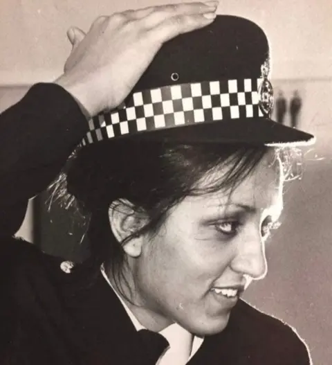 Anjum Malik Black and white photo of Anjum in her police uniform, she is smiling, looking to the side whith one had on her head. 