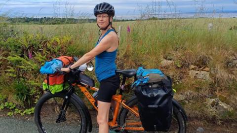 Becky Sherwood wearing a blue top and a helmet. She is standing with her orange bike next to the countryside
