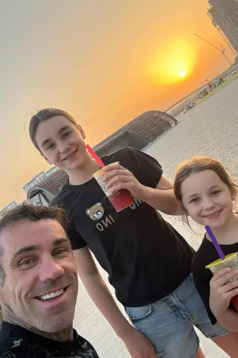 Ryan Hodgson Ryan Hodgson smiles next to his two daughters, They are all wearing black tshirts. The sun is setting behind them. 