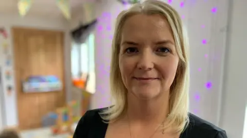 BBC Theresa McCaughey has shoulder-length blonde hair and is looking at the camera. She has a gold necklace around her neck and is wearing a dark coloured top. There are children's toys in the background
