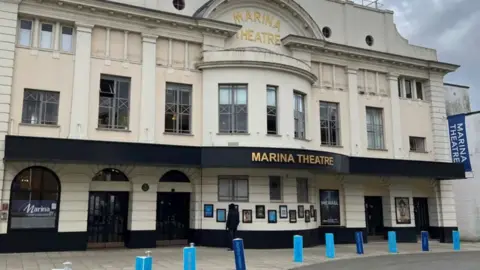 Richard Knights/BBC An image of the outside of the Marina Theatre in Lowestoft taken from the road in front of it. It is a large cream-coloured and grand-looking building with posters on the lowest walls either side of the entrance doors. 