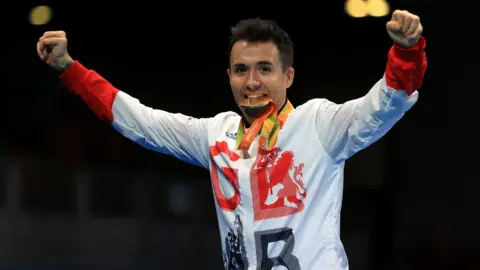 PA Media Will Bayley, wearing a GB sports shirt as he bites a gold medal. Both his arms are stretched out to the side in victory.