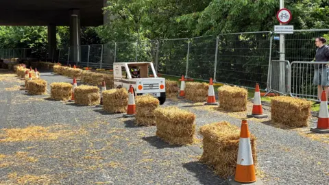 A person wearing a black helmet inside of a small homemade kart riding between a line of straw bales and orange cones. 
