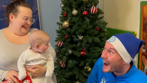 John Devine/BBC Baby Luna-Violet is in a white baby grow and is being held by her mum. She is looking at Jed Steer who wears a blue Peterborough FC branded Santa hat and is entertaining Luna-Violet by making faces. His mouth and eyes are open wide and they are both smiling. In the background there is a Christmas tree on the hospital ward. 