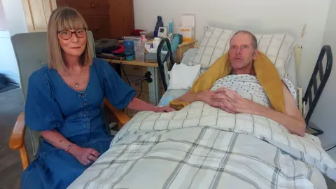 Soo and Tim Hall. Sue has a straight blond hair along the shoulder and wears a blue dress and glasses. She sits next to Tim, who is in bed. He has his arms folded on his bosom and a yellow blanket around his neck and shoulders.