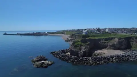 Seaham Town Council A view of the coastline at Seaham and some of the houses of the town. The water is a deep blue and it is sunny.