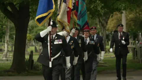 Five standard bearers, fully dressed in military uniforms, march in a cemetery in Hull. 