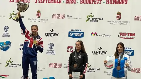 Freya Holmes Freya Holmes is stood on the winning podium. She is wearing a red, white and blue GB tracksuit and is holding a large gold cup aloft in one hand. In the other hand, she is holding a large gold medal. Two women are stood next to her in tracksuits, both with medals and certificates