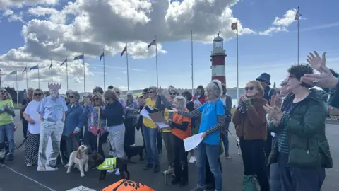 A crowd of people stood together singing. There is a cardboard cut out of Sir David Attenborough on the left. 