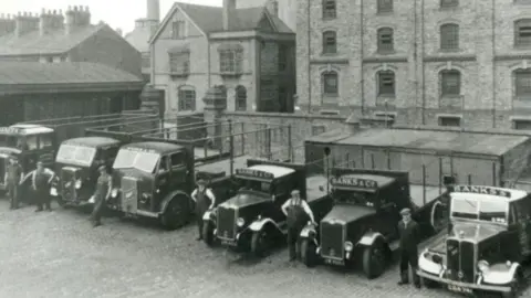 Carlsberg Britvic Group A black and white photo of a row of lorries with people in suits and hats standing in front of them and a large brick building with several floors in the background