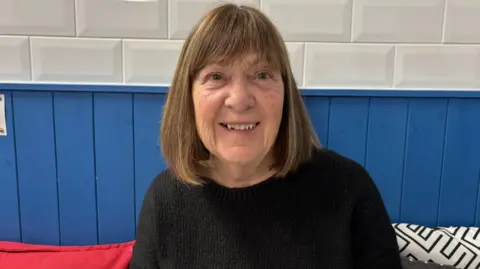 A woman wearing a black jumper smiles a the camera. She is sitting on a bench in a cafe. Blue wooden cladding is behind her.