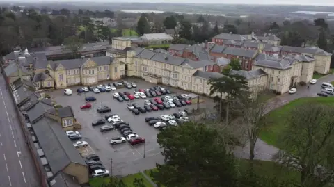 Dawid Wojtowicz Aerial pictures of a large hospital site. There are numerous buildings which are attached or contiguous to one another. The buildings are coloured off white or a shade of orange-red. There is a car park. To the left of the picture is a busy road.