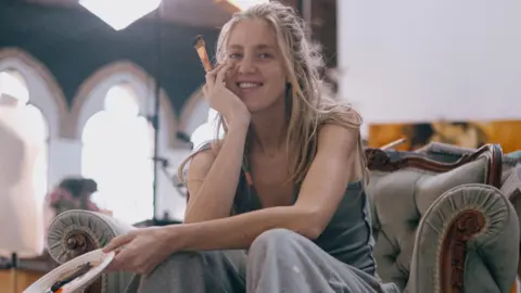 A woman with long blonde hair, wearing grey trousers and a vest top, sits on an armchair holding a plate palette and a brush