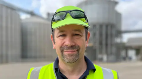 Tom Jackson/BBC A man - Simon Willis - looking straight at the camera. He is standing in front of several silos and is wearing a yellow high visibility vest and matching hat.