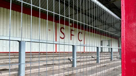 A metal fence blocks entry to a football stand. On the wall are the initials SFC in red lettering