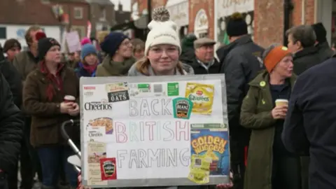 A young woman holds up a sign that reads "back British farming". Several logos of well-known brands are glued to the sign such as Heinz, Cheerios and Quavers crisps. She stands in the middle of the market square, surrounded by others who joined the rally.