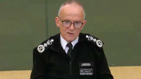 London Policing Board Sir Mark Rowley in black police uniform. He is sitting behind a desk at City Hall and has short grey hair and glasses.